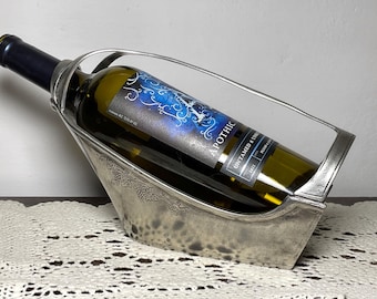 May include: A silver wine bottle holder with a decorative grape vine design. The holder cradles a bottle of white wine with a blue and white label that reads "Apothic Untamed & Uncorked". The bottle is angled in the holder.