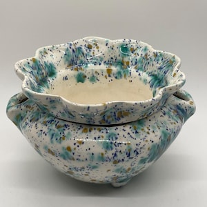May include: A ceramic planter with a scalloped rim and a rounded base, featuring a white glaze decorated with a speckled pattern of teal, blue, and mustard yellow paint splatters. The vessel has a wide opening and a distinct two-part design.