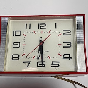 May include: A vintage General Electric alarm clock with a red frame and silver accents. The clock face is white with black numerals and red hands. The clock is rectangular and has a power cord.