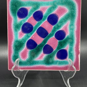 May include: A square ceramic tile with a pink and green background and blue circles. The tile has a geometric pattern.
