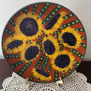 May include: A round, hand-painted ceramic plate featuring a sunflower design. The plate has a dark orange background with yellow sunflowers, green accents, and a green rim. The sunflowers have purple centers and yellow petals.