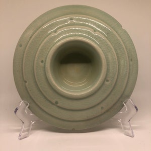 May include: A light green ceramic bowl with a raised center and concentric circles around the rim. The bowl has a textured surface and several small holes around the rim.