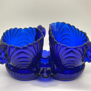 May include: A pair of cobalt blue glass sugar bowls with matching saucers, connected by a central handle. The bowls have a scalloped rim and a textured, fan-like design. Each bowl has a small handle on the side.