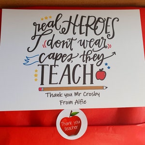 May include: A white card with a red apple and the text "real HEROES (I don't wear capes, they TEACH)". The card also has the text "Thank you Mr Crosby From Alfie" and a red apple sticker with the text "Thank you teacher".