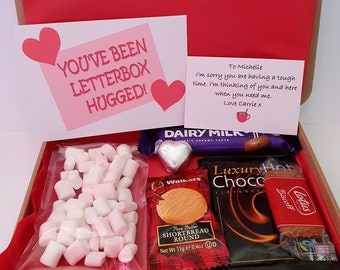 May include: A red and white gift box filled with treats, including chocolate bars, cookies, marshmallows, and a handwritten card that says "You've been letterbox hugged!" and "To Michelle, I'm sorry you are having a tough time. I'm thinking of you and here when you need me. Love Carrie x."
