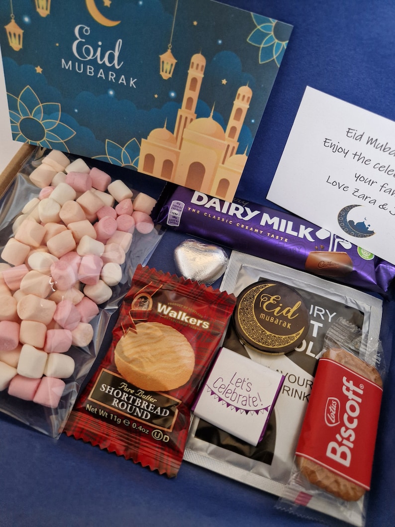 May include: A gift box filled with treats for Eid Mubarak. The box includes a chocolate bar, shortbread cookies, marshmallows, and a small card with a mosque illustration. The card reads "Eid Mubarak".