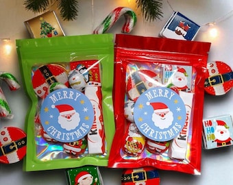 May include: Two Christmas-themed gift bags, one green and one red, filled with candies and treats. Each bag has a blue circular sticker that says "Merry Christmas" with a Santa Claus graphic. Candy canes and small gift boxes are scattered around.