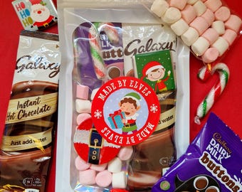 May include: A festive assortment of treats on a red background. Includes a bag of Galaxy instant hot chocolate, marshmallows, candy canes, and a Cadbury Dairy Milk Buttons bag. The bag is labeled "Made by Elves" with a cartoon elf.