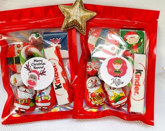 May include: Two red cellophane bags filled with Christmas candies and treats. Each bag contains a variety of sweets, including candy canes, chocolate Santas, and Kinder bars. One bag has a sticker that says "Merry Christmas Nanna!" and the other says "Millie".