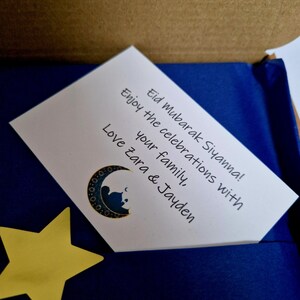 May include: A white card with a blue background and a crescent moon and star design. The card reads "Eid Mubarak Siyanna! Enjoy the celebrations with your family, Love Zara & Jayden".