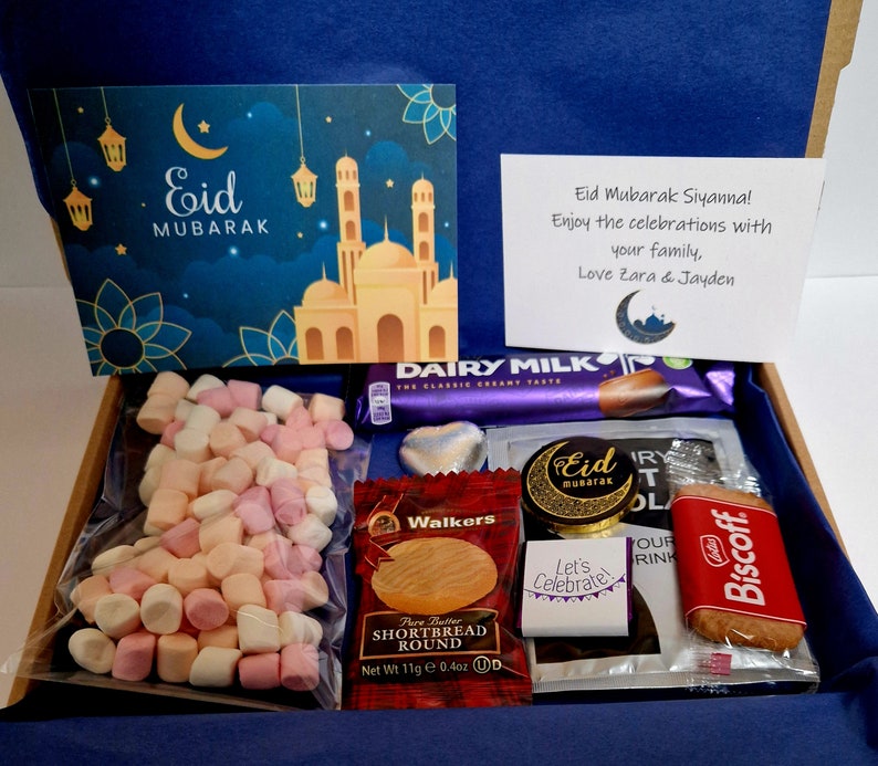 May include: A gift box filled with treats for Eid Mubarak. The box includes a card with a mosque illustration, a chocolate bar, marshmallows, shortbread cookies, and a Biscoff cookie.