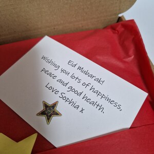 May include: A white card with a handwritten message wishing someone a happy Eid Mubarak. The message includes wishes for peace, happiness, and good health. The card is decorated with a gold star.