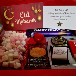 May include: A red gift box filled with various treats for Eid Mubarak. The box contains a chocolate bar, shortbread cookies, biscuits, marshmallows, and a card with a message wishing happiness, peace, and good health.