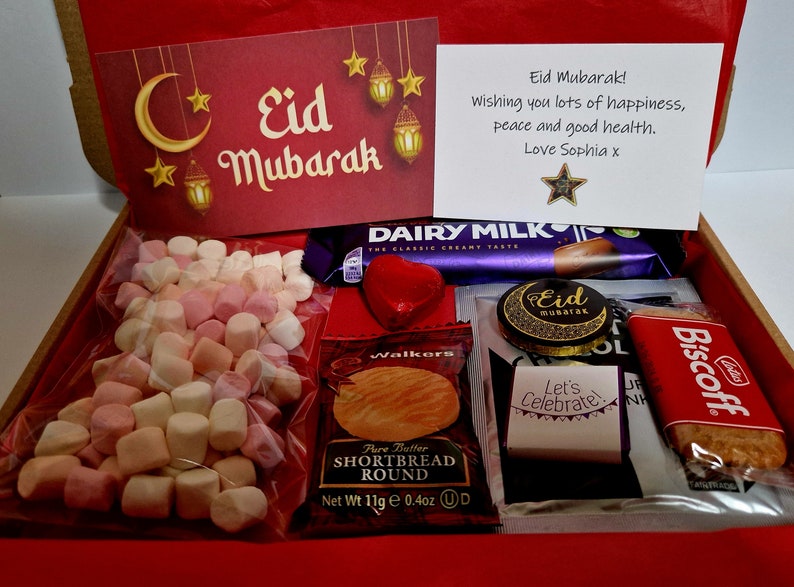 May include: A red gift box filled with various treats for Eid Mubarak. The box contains a chocolate bar, shortbread cookies, biscuits, marshmallows, and a card with a message wishing happiness, peace, and good health.