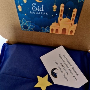 May include: A blue gift box with a card that says "Eid Mubarak Siyanna! Enjoy the celebrations with your family, Love Zara & Jayden". The box has a sticker on the top with a mosque and the words "Eid Mubarak" in gold lettering.