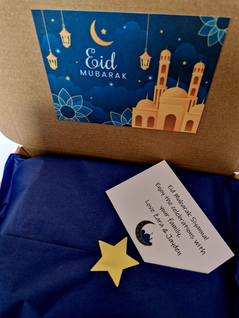 May include: A blue gift box with a card that says "Eid Mubarak Siyanna! Enjoy the celebrations with your family, Love Zara & Jayden". The box has a sticker on the top with a mosque and the words "Eid Mubarak" in gold lettering.