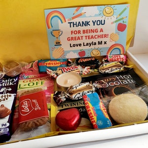 May include: A white gift box filled with various chocolate bars, candies, biscuits, and a small candle. The box is decorated with a yellow card that says "Thank you for being a great teacher! Love Layla Mx".