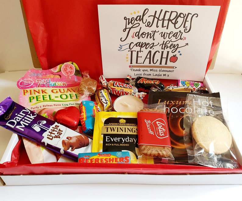 May include: A gift box filled with various treats, including chocolate bars, tea bags, biscuits, and a candle. The box is decorated with red tissue paper and a card that reads "real HEROES (don't wear) capes, they TEACH*" with a red apple emoji.