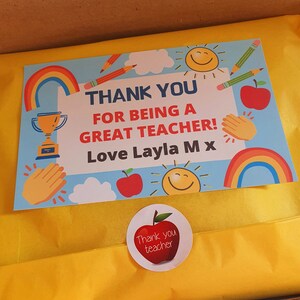 May include: A colorful card with a rainbow, a trophy, a sun, pencils, and apples. The card says "Thank you for being a great teacher! Love Layla Mx". There is a sticker on the yellow paper that says "Thank you teacher".