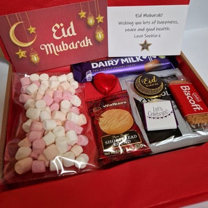 May include: A red gift box filled with treats for Eid Mubarak. The box contains a chocolate bar, a bag of marshmallows, a chocolate coin, a shortbread cookie, a biscuit, and a card with the message "Eid Mubarak! Wishing you lots of happiness, peace and good health. Love Sophia x".