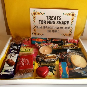 May include: A white gift box filled with various treats, including chocolate bars, biscuits, candy, and tea. The box is decorated with a card that reads "Treats for Mrs. Sharp. Thank you for helping me grow! Love Aisha X."