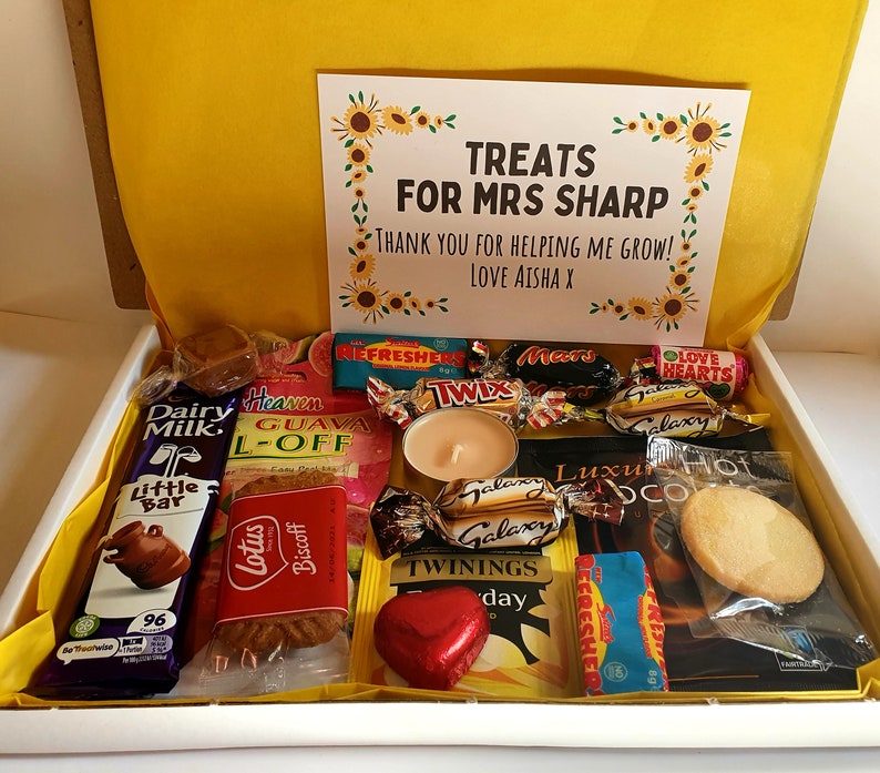 May include: A white gift box filled with various treats, including chocolate bars, biscuits, candy, and tea. The box is decorated with a card that reads "Treats for Mrs. Sharp. Thank you for helping me grow! Love Aisha X."