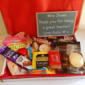 May include: A gift box filled with various treats, including chocolate bars, biscuits, tea bags, and a candle. The box is lined with red paper and has a handwritten card that reads "Mrs Jones Thank you for being a great teacher! Love Aisha M x".