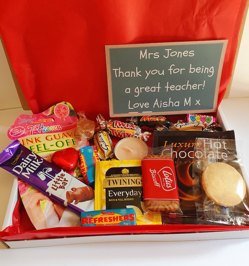 May include: A gift box filled with various treats, including chocolate bars, biscuits, tea bags, and a candle. The box is lined with red paper and has a handwritten card that reads "Mrs Jones Thank you for being a great teacher! Love Aisha M x".