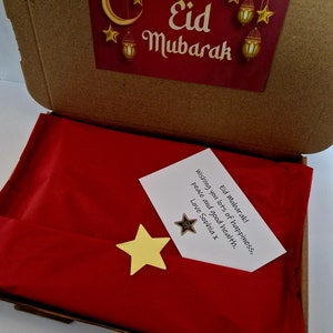 May include: A red gift box with a white card inside that says "Eid Mubarak! Wishing you lots of happiness, peace and good health. Love Sophia x". The box is decorated with a gold star and a crescent moon.