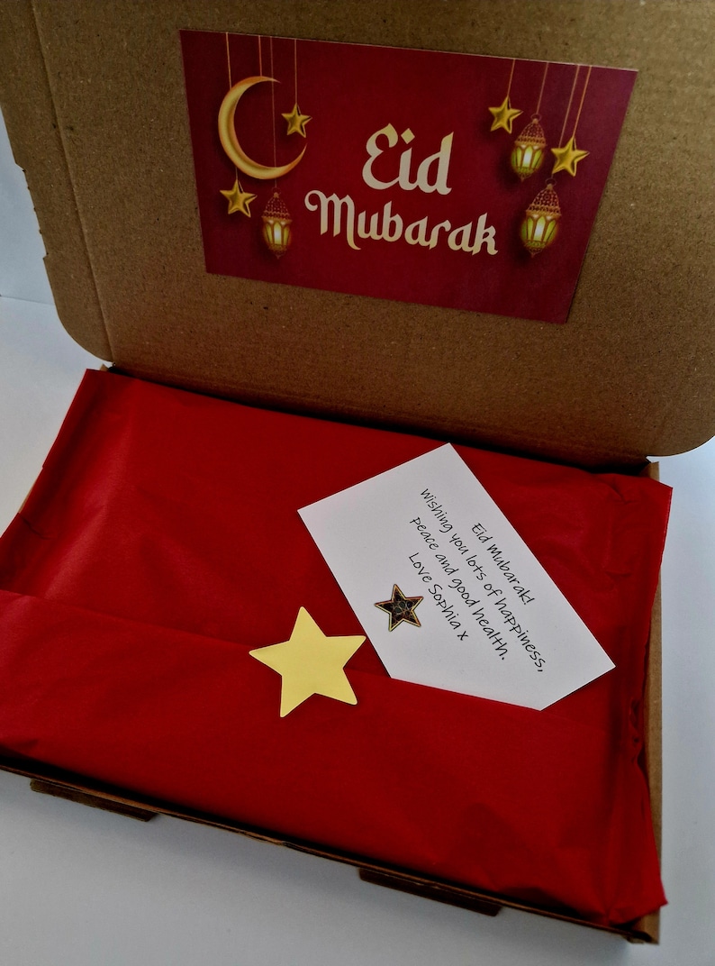 May include: A red gift box with a white card inside that says "Eid Mubarak! Wishing you lots of happiness, peace and good health. Love Sophia x". The box is decorated with a gold star and a crescent moon.