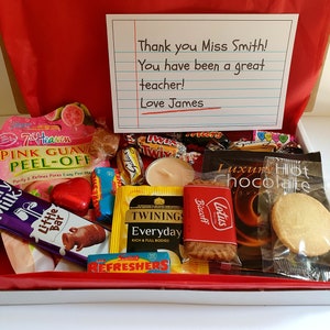 May include: A gift box filled with various treats, including chocolate bars, cookies, tea bags, and a candle. The box is lined with red tissue paper and has a handwritten note that reads "Thank you Miss Smith! You have been a great teacher! Love James".