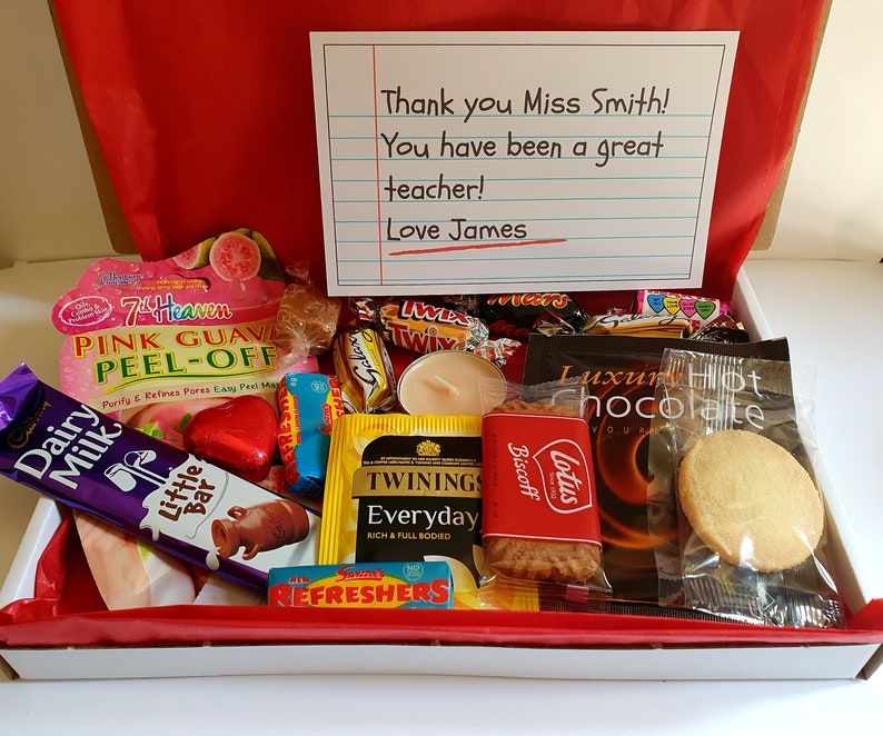 May include: A gift box filled with various treats, including chocolate bars, cookies, tea bags, and a candle. The box is lined with red tissue paper and has a handwritten note that reads "Thank you Miss Smith! You have been a great teacher! Love James".