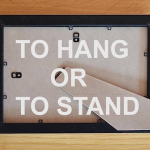 May include: Black picture frame with a brown backing. The backing has a fold-out stand and the text "TO HANG OR TO STAND" printed in white.