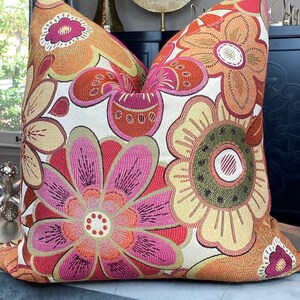 May include: A decorative cushion featuring a floral design in shades of pink, orange, yellow, and green. The cushion is made from a soft, textured fabric and has a white background.