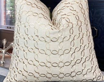 May include: A beige throw pillow with a gold geometric pattern. The pillow is sitting on a grey velvet ottoman.