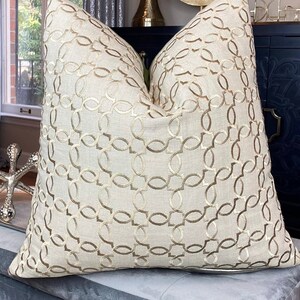 May include: A beige throw pillow with a gold geometric pattern. The pillow is sitting on a grey velvet ottoman.