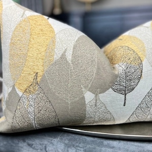 May include: A decorative throw pillow with a beige and gold leaf pattern. The pillow is sitting on a gray velvet surface.
