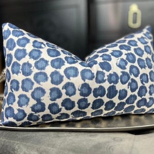 May include: A rectangular throw pillow featuring a repeating pattern of dark blue, circular, watercolor-style spots on a cream-colored background. The fabric has a visible woven texture, and the pillow is filled to create a plump, structured shape.