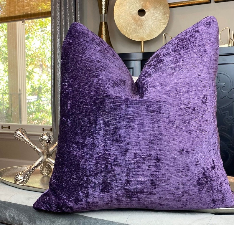 May include: A purple velvet cushion with a textured, slightly iridescent finish. The cushion is square and has a simple, elegant design.