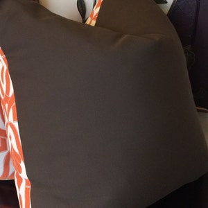 Pode incluir: Uma almofada marrom com uma borda estampada em laranja e branco. A almofada est&aacute; sentada em uma superf&iacute;cie branca com uma moldura de madeira marrom.