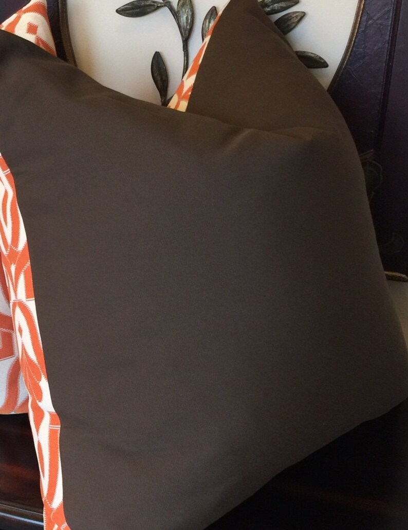 Pode incluir: Uma almofada marrom com uma borda estampada em laranja e branco. A almofada est&aacute; sentada em uma superf&iacute;cie branca com uma moldura de madeira marrom.
