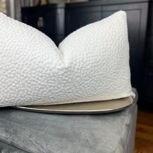 May include: A white, textured cushion with a subtle raised pattern. The cushion is sitting on a grey ottoman.
