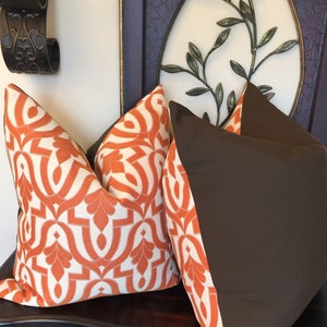 Pode incluir: Duas almofadas decorativas, uma com tecido marrom e a outra com um padr&atilde;o geom&eacute;trico branco e laranja.