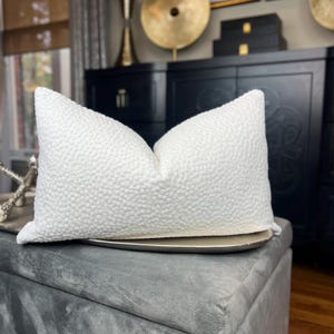 May include: White textured cushion with a subtle pattern. The cushion is sitting on a grey ottoman.