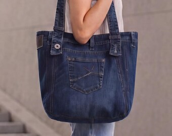 May include: A dark blue denim tote bag constructed from repurposed jeans material, featuring a visible back pocket, belt loops, a metal button detail, and two wide shoulder straps. The bag has a structured, rectangular shape with reinforced stitching.