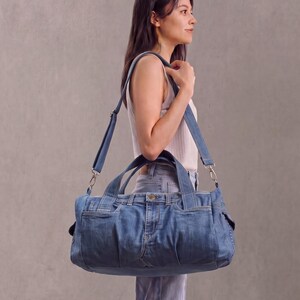 Könnte beinhalten: Eine mittelblaue Reisetasche aus Denim, gefertigt aus wiederverwendeten Jeans, mit einem Reißverschluss an der Oberseite, zwei kurzen Tragegriffen und einem verstellbaren Schultergurt mit silberfarbenen Metallclips an den Seiten.