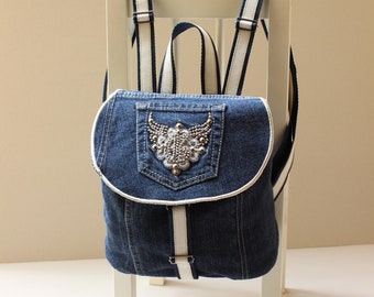 May include: A blue denim backpack with white straps and a decorative embellishment on the front pocket.