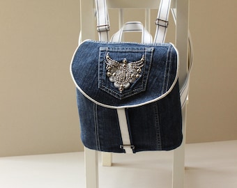 May include: A blue denim backpack with white straps and a decorative embellishment on the front pocket.