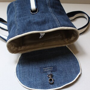 May include: A blue denim backpack with a white trim and a white strap. The backpack has a zipper closure and a small pocket on the front. The backpack is open and the inside is lined with a beige fabric. The backpack has a metal hook on the inside.