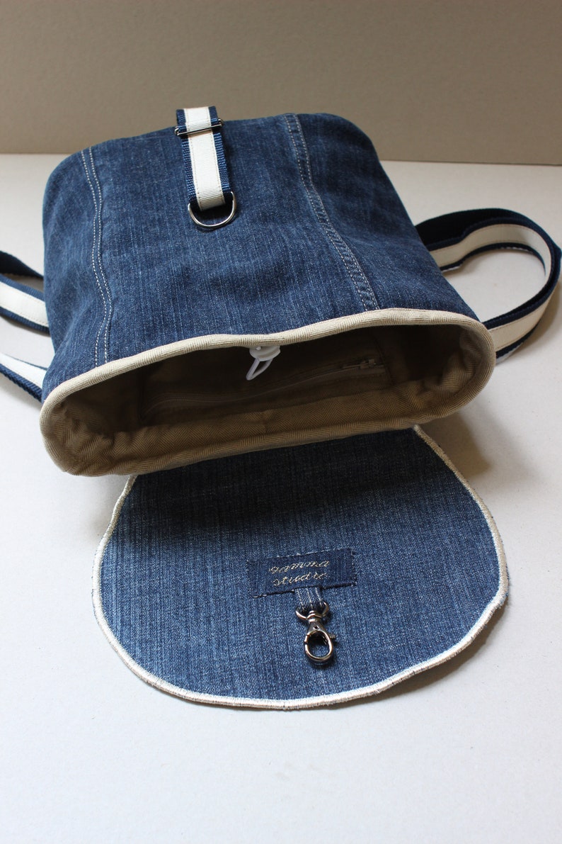 May include: A blue denim backpack with a white trim and a white strap. The backpack has a zipper closure and a small pocket on the front. The backpack is open and the inside is lined with a beige fabric. The backpack has a metal hook on the inside.
